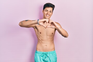 Fototapeta premium Young hispanic man wearing swimwear shirtless smiling in love doing heart symbol shape with hands. romantic concept.