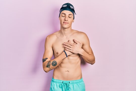 Young hispanic man wearing swimwear and swimmer glasses smiling with hands on chest with closed eyes and grateful gesture on face. health concept.