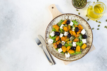 Healthy salad with fried pumpkin, feta cheese, olives, salad greens, pumpkin seeds, olive oil on the light grey background.