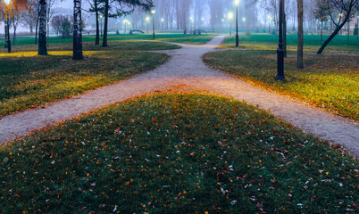 Different trails, routes converge and diverge in different directions in the foggy twilight and the light of night lanterns in the park in autumn.