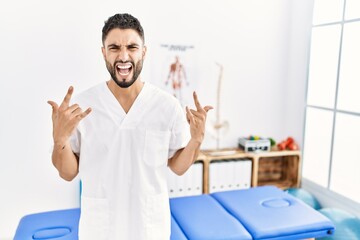 Young handsome man with beard working at pain recovery clinic shouting with crazy expression doing rock symbol with hands up. music star. heavy concept.
