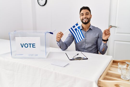Young handsome man with beard at political campaign election holding greece flag smiling happy pointing with hand and finger to the side