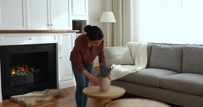 Making home cozy. Happy young Indian lady put bunch of lavender flowers on small table after cleaning living room. Caring millennial housewife decorate flat enjoy comfort at modern luxury apartment