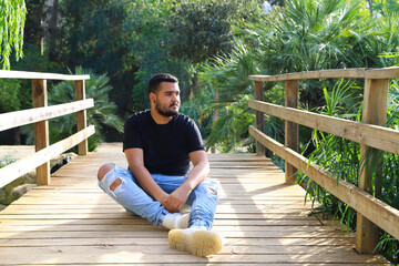 A gay man of Latin origin, dark and bearded, stands pensively looking nowhere in the park. He is...