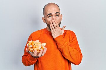 Young bald man holding potato chip covering mouth with hand, shocked and afraid for mistake....
