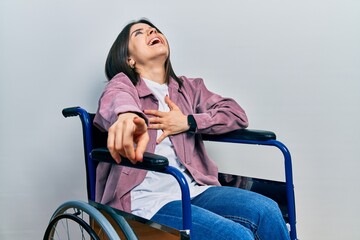 Obraz premium Young brunette woman sitting on wheelchair laughing at you, pointing finger to the camera with hand over body, shame expression