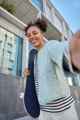 Young attractive teenage girl poses for making selfie holds skateboard enjoys recreation time in urban setting dressed in street style clothes smiles happily. Youth and everyday life concept © WHstudio Leushin N