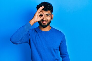 Arab man with beard wearing casual blue sweater doing ok gesture with hand smiling, eye looking through fingers with happy face.