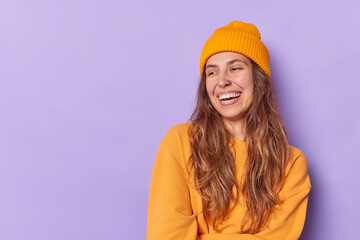 Studio shot of happy teenage girl with toothy smile laughs joyfully has piercing in nose wears orange hat and sweatshirt isolated over purple background blank space for your promotional content