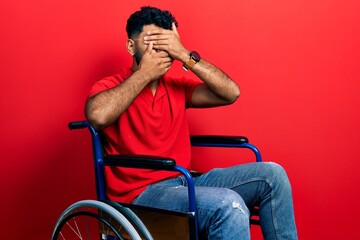 Arab man with beard sitting on wheelchair covering eyes and mouth with hands, surprised and shocked. hiding emotion