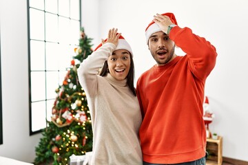 Young hispanic couple standing by christmas tree surprised with hand on head for mistake, remember error. forgot, bad memory concept.