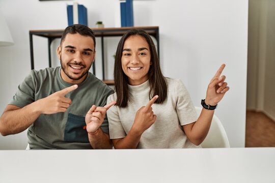 Young latin couple wearing casual clothes sitting on the table smiling and looking at the camera pointing with two hands and fingers to the side.