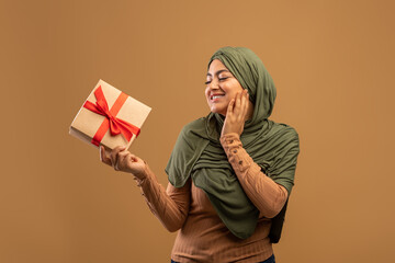 Excited arab young lady in hijab holding gift box, wondering what is inside, standing over brown...