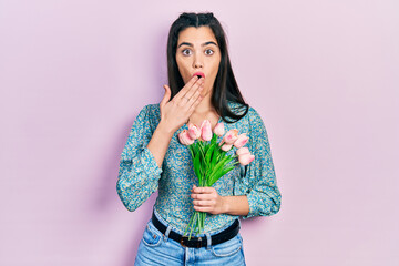 Young hispanic girl holding flowers covering mouth with hand, shocked and afraid for mistake....