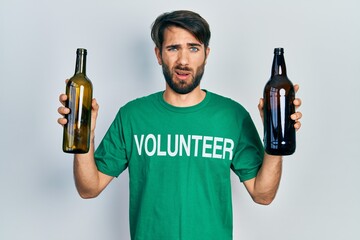 Young hispanic man wearing volunteer t shirt holding recycling bottle glass in shock face, looking...