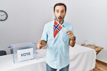 Middle age man with beard at political campaign election holding norway flag scared and amazed with...