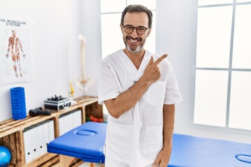 Middle age man with beard working at pain recovery clinic cheerful with a smile of face pointing with hand and finger up to the side with happy and natural expression on face