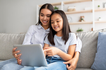 Excited asian mother and daughter using digital tablet together while sitting on sofa and surfing internet, free space