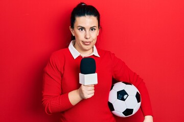 Young hispanic woman holding reporter microphone and soccer ball relaxed with serious expression on...