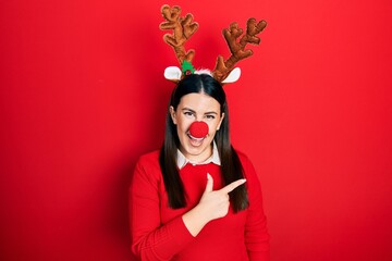 Young hispanic woman wearing deer christmas hat and red nose cheerful with a smile on face pointing with hand and finger up to the side with happy and natural expression