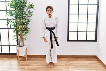 Young hispanic girl wearing karate kimono and black belt afraid and shocked with surprise...