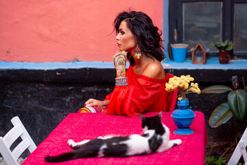 Beautiful woman with curly hair and makeup in sensual dress, sitting at the table drinking a coffee, looking at the cat.