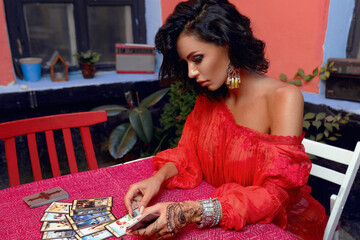 Beautiful woman with curly hair and makeup in sensual dress, sitting at the table arrange tarot cards. Horizontal view.
