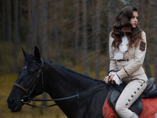 Obraz premium A young beautiful brunette rider in an elegant retro suit riding a black horse, in a forest area