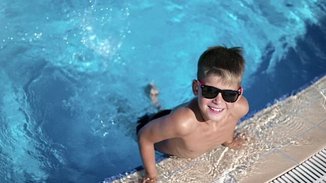 Portrait baby boy in trendy sunglasses playing splash water at swimming pool summer vacation slowmo