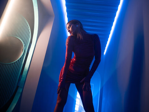 A fashionable photo shoot with tinted blue, a young beautiful model in a scarlet dress, in a mysterious corridor with smoke and club lighting