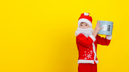 Fototapeta na wymiar A cute Caucasian child in Santa Claus clothes and with an artificial white beard holds a round gift box high in his hands with the words New Year 2022, yellow studio background.