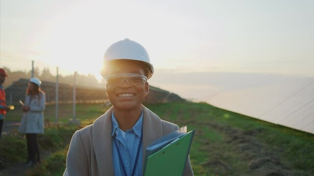Young african professional female engineer wearing glasses and helmet smiling cheerful outdoors. Nature conservation. Sustainable solar station. Ecology. Portraits.