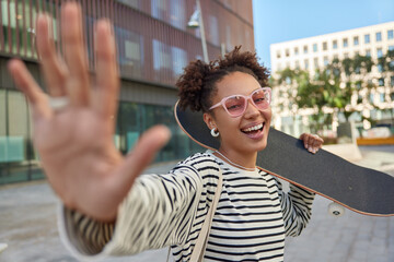 Carefree cheerful woman keeps palm forward camera smiles positively enjoys riding skateboard wears...