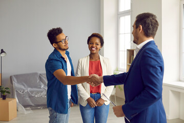 Obraz premium New house, new beginnings. Happy young married couple buys house and closes deal with realtor with handshake. African American family gratefully shakes hands with Caucasian real estate agent.