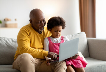 Happy african american small girl and grandfather surfing in internet or playing an online game on laptop