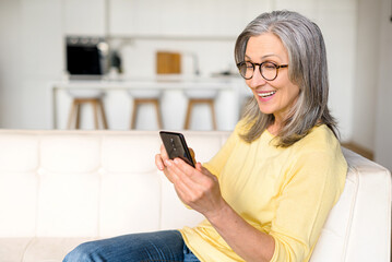 Positive senior mature lady sitting in a relaxed pose on the comfortable couch with a smartphone, an older gray-haired woman spends leisure time scrolling news feed, web surfing