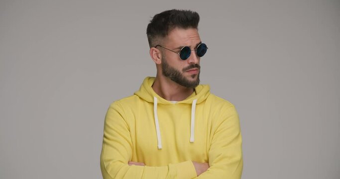 cool bearded man with retro sunglasses in yellow hoodie crossing arms, looking to side and nodding on grey background in studio