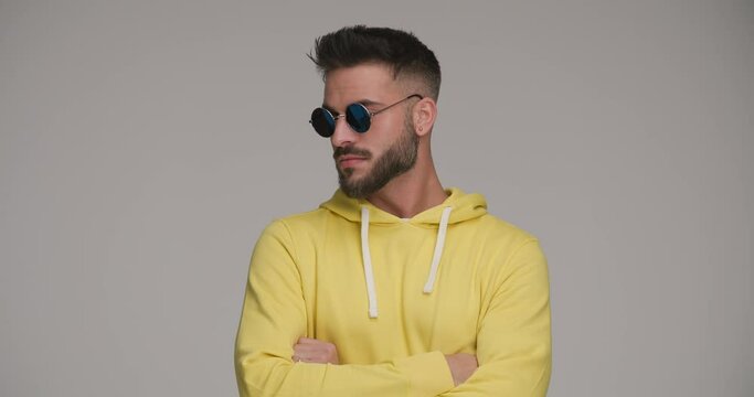 handsome bearded man with retro sunglasses folding arms, looking to side, pointing fingers, smiling and applauding in front of grey background