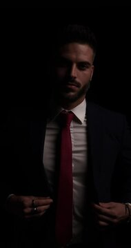 mysterious bearded man in black suit buttoning jacket, looking to side and rubbing palms in a sexy manner on dark background in studio