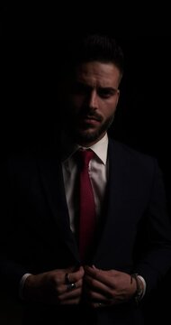 vertical video of attractive young businessman buttoning suit, fixing tie and looking to side, moving in a side view position and fading in the dark