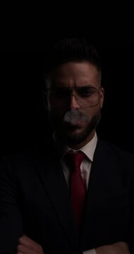 attractive unshaved young man crossing arms, smoking and looking to side in front of black background