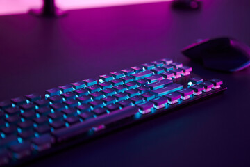 Computer mouse and illuminated keyboard laying on desk. Pink light in background.