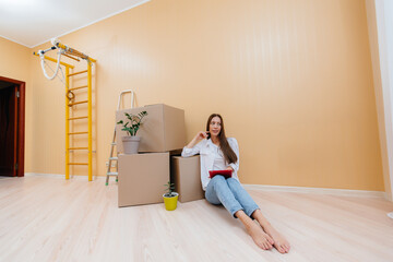A young woman is sitting on the floor in front of the boxes and picks up a new design for her apartment on a tablet after moving. Housewarming, delivery and cargo transportation.