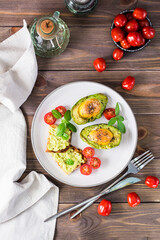 Hot appetizer baked avocado with egg, toast, cherry tomatoes and basil on a plate on a wooden table. Healthy eating. Flexitarian diet. Top and vertical view