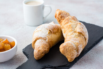 A couple of fresh crispy croissants on a slate, a cup of coffee and jam in a bowl on a light table. Coffee break or breakfast