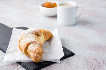 Fresh crispy croissant in powdered sugar on paper on a slate, a cup of coffee and jam in a bowl on a light table. Coffee break or breakfast