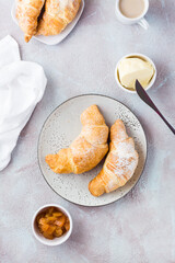 Fresh crispy croissants on plates, a cup of coffee and bowls with jam and butter on a light textured background. Coffee break or breakfast. Vertical and top view