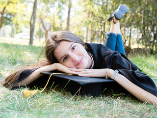 Portrait of a happy smiling girl in a black robe lying on the grass. A young woman is celebrating her graduation. New life