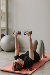 Young pregnant woman doing exercises with dumbbells laying on mat in living room