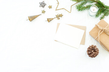 Christmas decorations composition.Christmas greeting card mockup scene. Blank greeting card and envelope,  Christmas box, fir branch, pine cone, decorations and burning candle.White linen background.